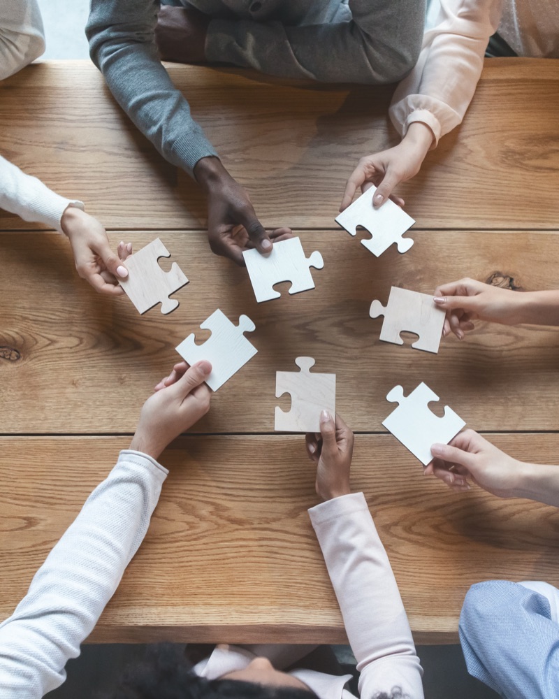 Diverse team members around a table, each holding a puzzle piece — every person brings a unique strength
